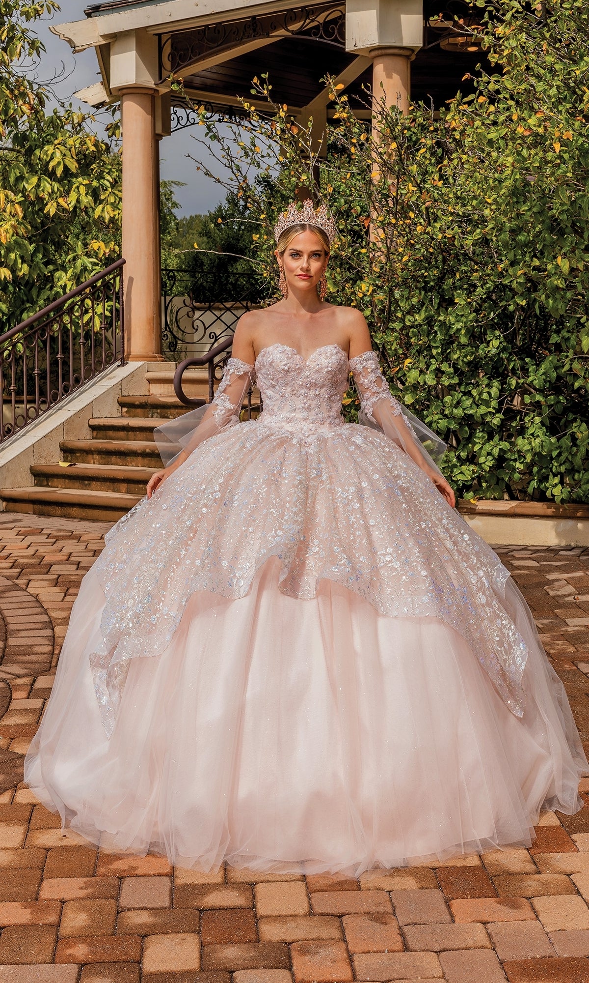 A woman in the Quinceanera Dress 1773 By Dancing Queen—sparkling, light pink, strapless with removable sheer sleeves and a tiara—stands outdoors on a paved walkway before steps and greenery.
